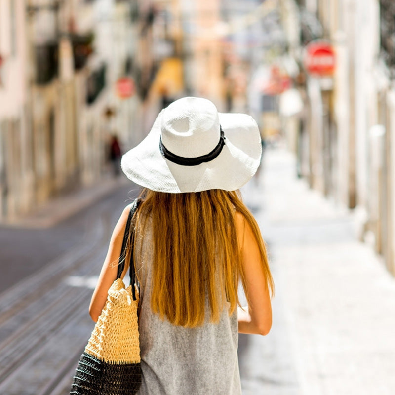 Woman wearing a white sun hat and carrying a straw bag on a city street.