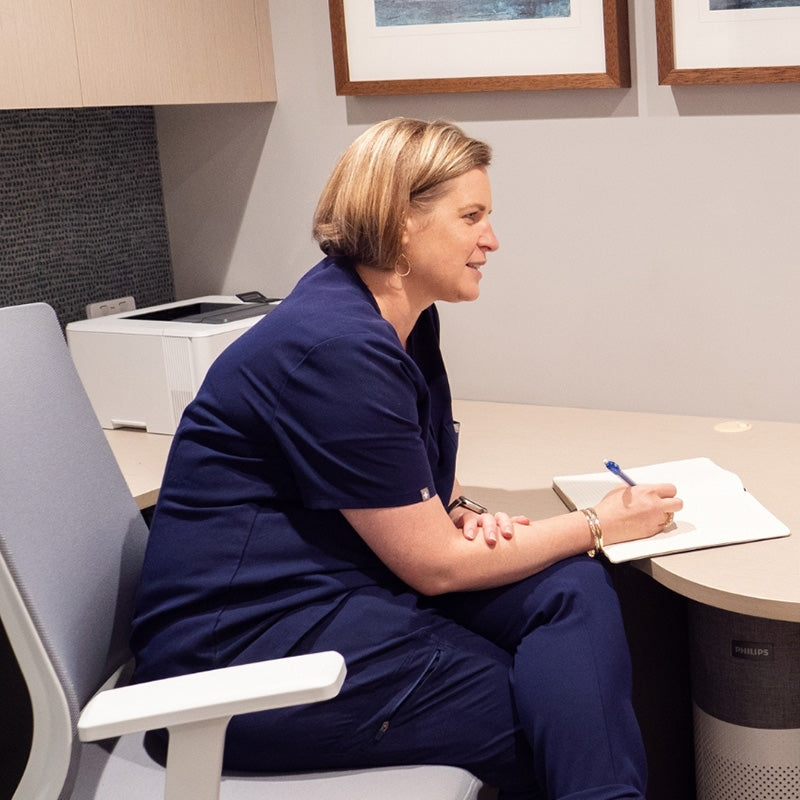 Woman in a blue uniform sitting at a desk with a notepad and pen, in an office setting.